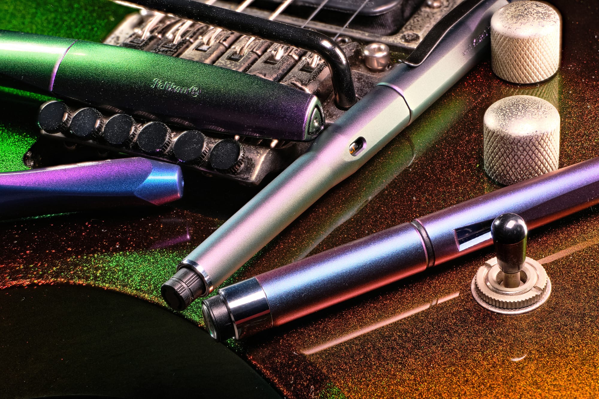 Photo of 3 fountain pens & a mechanical pencil sitting on top of the body of an electric guitar. All items have different colour-shifting paint jobs.