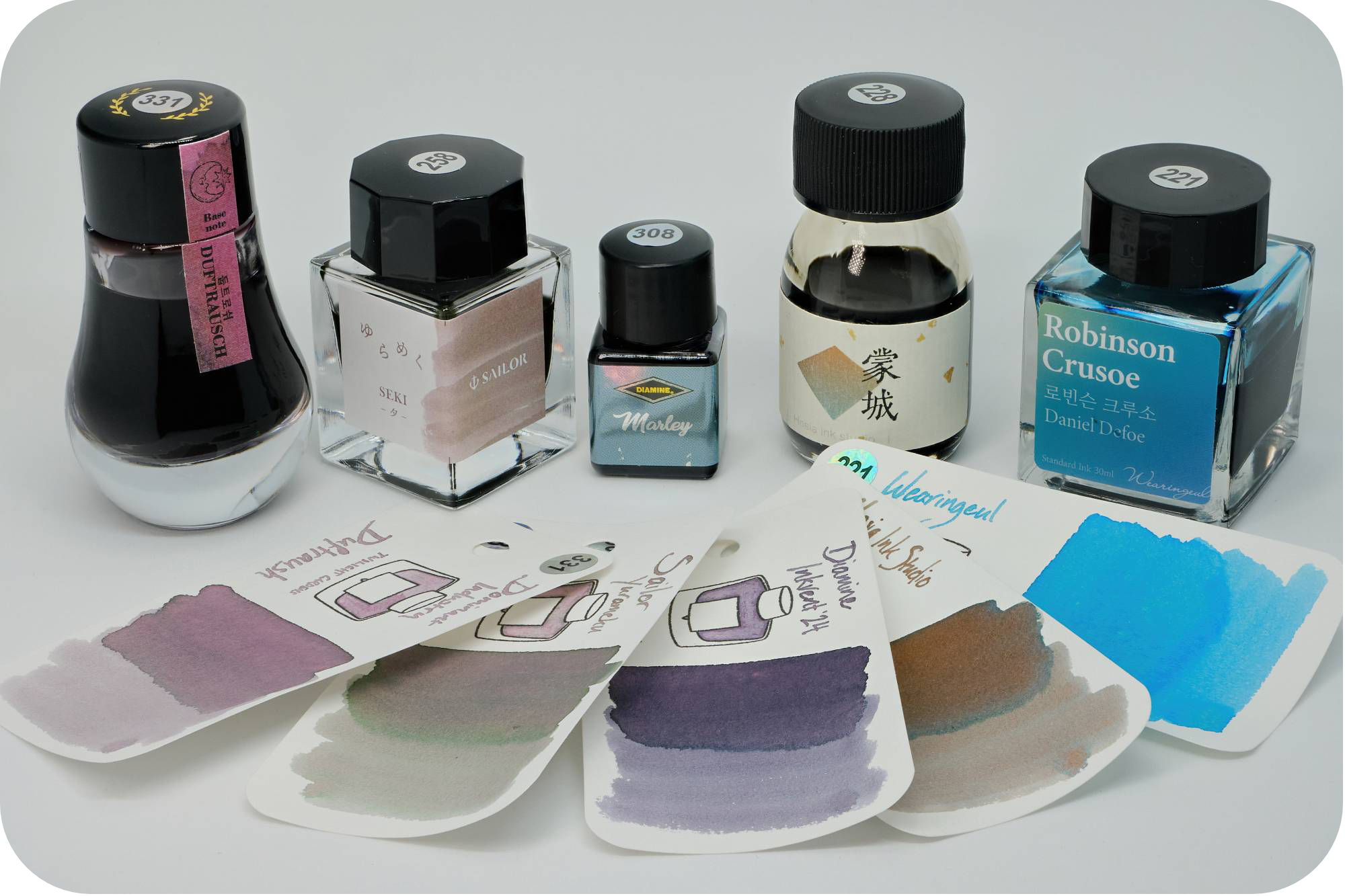 A photo of Dominant Industry Duftraush, Sailor Seki, Diamine Marley, Hosia Ink Studio Mengchen, & Wearingeul Robinson Crusoe bottles behind their respective swatches on a white background