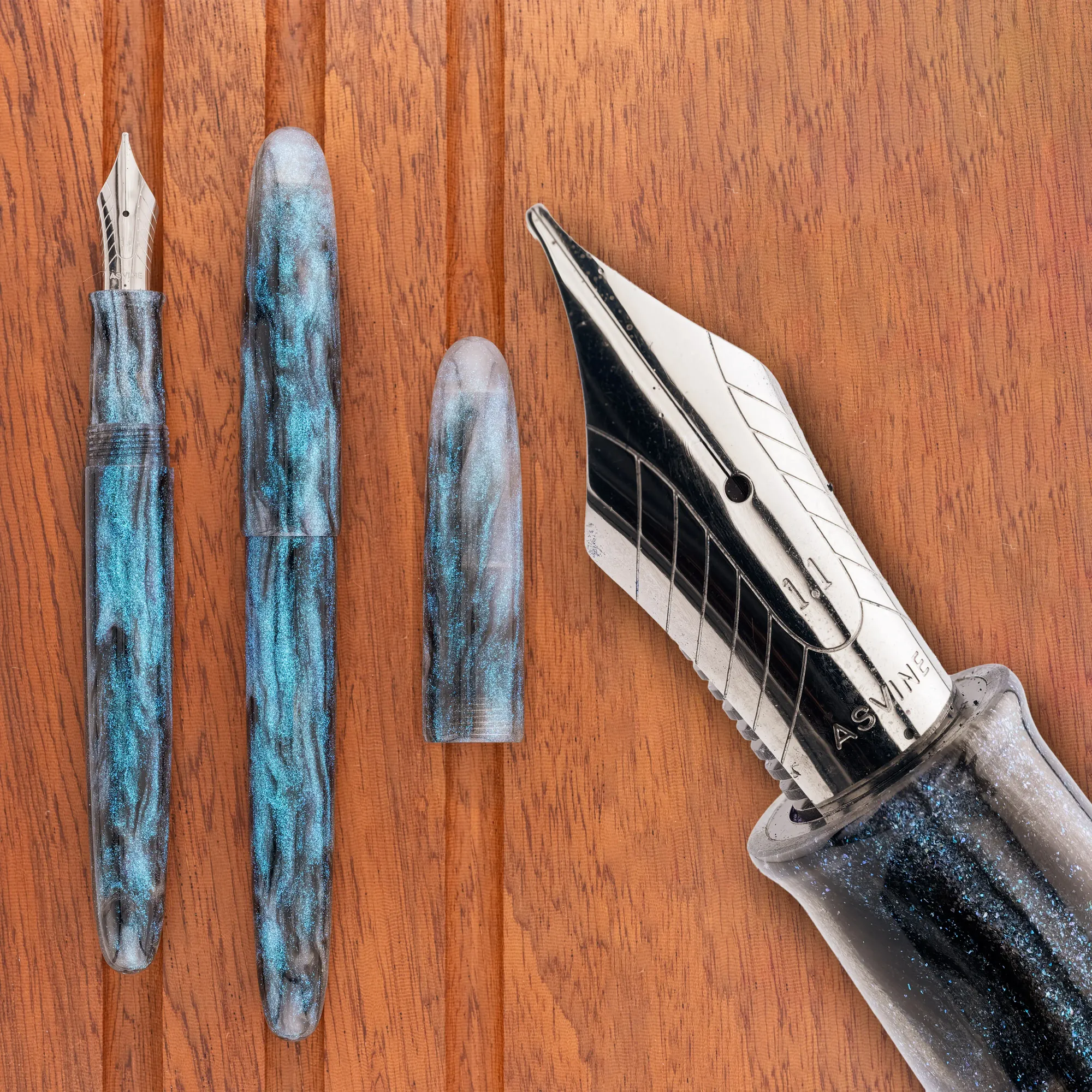 Overhead photo of a blue, grey, & white swirly acrylic with blue sparkles, torpedo-shaped fountain pen on a wooden tray. The pen is shown capped and uncapped, along with a close-up of the nib.