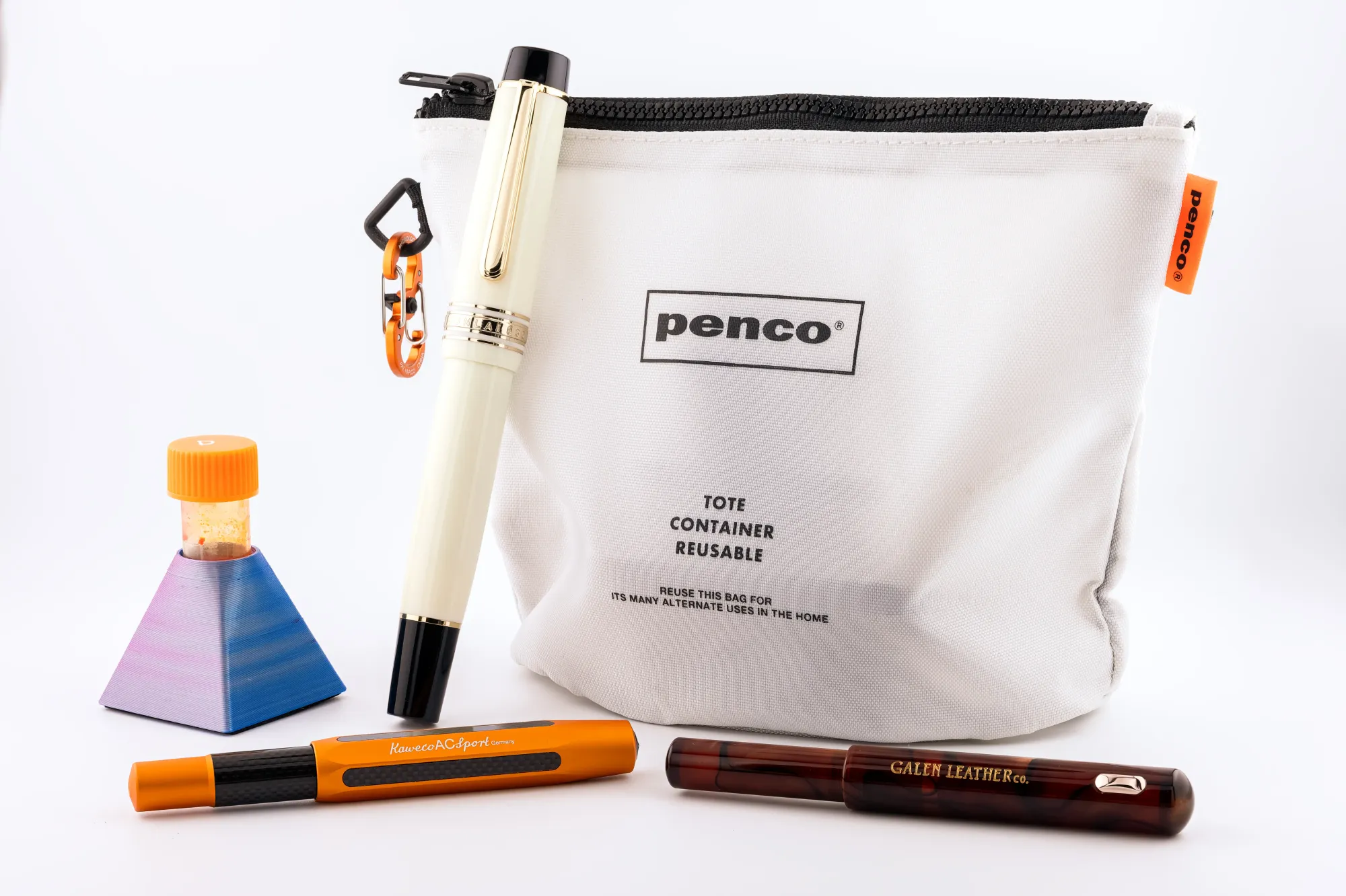 Photo of an orange-capped 5ml sample vial in a blue/purple pyramid-shaped holder, an ivory & black Jun Lai 930 resting on a white Penco Bucket Pouch, with an orange Kaweco AC Sport and brown Fine Writing International × Galen Leather Pencket in front, all on a white background.